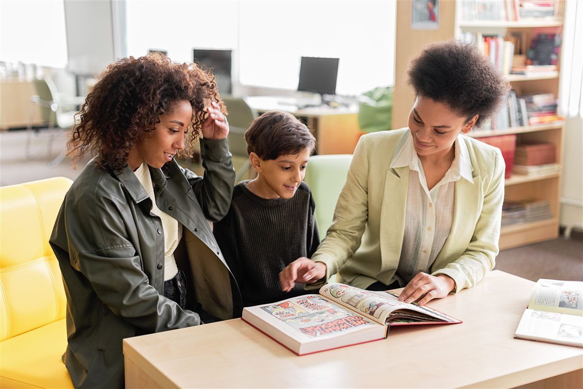 A tutor reading a book with a young boy and his mother at a library table